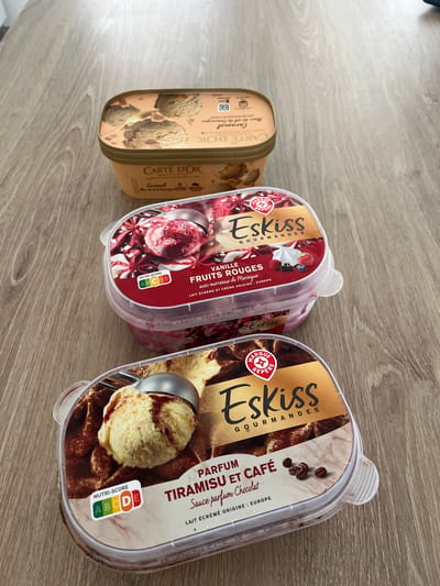 Three containers of frozen desserts are displayed on a light-colored wooden surface, likely a table or floor, in an indoor setting in Saint-Just-Saint-Rambert, France. The containers are positioned at an angle.

On the left is a rectangular carton of "CARTE D'OR Caramel fleur de sel de Camargue" ice cream. The label shows an image of ice cream scoops.

In the middle and on the right are two clear plastic tubs of "Eskiss GOURMANDES" frozen desserts. Both Eskiss tubs feature "MARQUE REPERE" (Repere Brand) and have images of ice cream scoops on their labels.
The middle tub is "Eskiss GOURMANDES VANILLE FRUITS ROUGES avec morceaux de Meringue" (Vanilla Red Fruits with Meringue pieces), with "LAIT ÉCRÉMÉ ET CRÈME ORIGINE : EUROPE" (Skimmed Milk and Cream Origin: Europe) stated. Its Nutri-Score is visible as C, highlighted in yellow/orange. An expiry or batch number "02/2028 15991 14-18 CB2" is also visible.
The right tub is "Eskiss GOURMANDES PARFUM TIRAMISU ET CAFÉ Sauce parfum Chocolat" (Tiramisu and Coffee flavor with Chocolate flavored sauce), also stating "LAIT ÉCRÉMÉ ORIGINE : EUROPE". Its Nutri-Score is visible as D, highlighted in orange.

The wooden surface exhibits a subtle grain pattern, and the lighting is bright and even. No human activity or other subjects are visible.