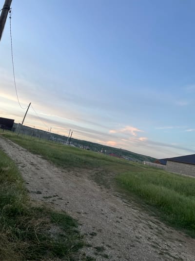 The image depicts an exterior scene in a rural or semi-rural area, possibly on the outskirts of Buynaksk, Russia, as suggested by the context. The sky is a clear, pale blue with some wispy clouds tinged with pink and orange, indicating it may be either early morning or late evening.
In the foreground, a dirt path, appearing to be a road, is visible, leading into the distance. The path is uneven and covered with gravel and some dry grass. To the left of the path, tall, green grass grows, with some dry straw-like vegetation mixed in.
Further into the scene, a wooden utility pole with a wire running from it is prominent on the left side of the frame. The wire extends diagonally across the image. Beyond the pole, a fence constructed of metal posts and wire mesh encloses an area. Several more utility poles with wires are visible in the middle ground, suggesting infrastructure in the area.
On the right side of the image, a building with a dark, sloped roof and a facade made of light-colored brick or concrete blocks is visible. A fence made of similar materials to the one on the left is also present. In the background, a gently sloping hill or range of hills is covered with green vegetation, with some scattered buildings or houses visible on its slopes. The overall impression is one of a developing residential area or a rural landscape with ongoing construction. There are no people or animals visible in the image.
