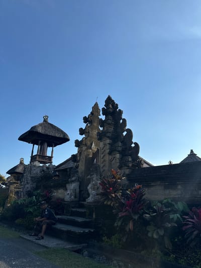 The image captures a traditional Balinese temple entrance, specifically located in Bangli, Indonesia. The architecture is characterized by intricate stone carvings and tiered roofs, common in Balinese religious structures. A prominent feature is a pavilion-like structure with a thatched roof, topped with a decorative crown. In the foreground, a man is seated on stone steps leading up to the temple complex. He is wearing traditional Balinese attire. The surrounding environment includes lush tropical foliage, with plants displaying vibrant red and green hues. The sky is clear and blue, suggesting a sunny day.
