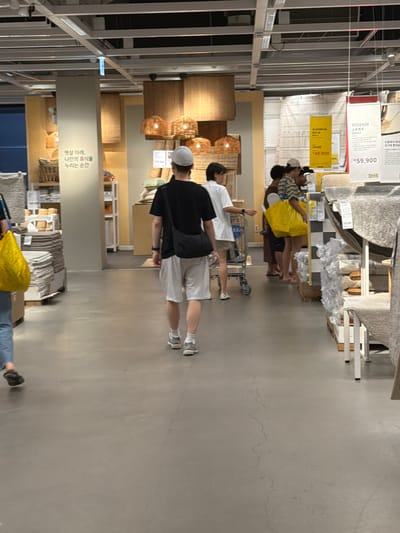 In Goyang-si, South Korea, this image captures the interior of an IKEA store, focusing on a section displaying home goods. The foreground shows the concrete floor, with several individuals walking through the aisle. To the left, a person in blue jeans and dark shoes walks away from the camera, carrying a large yellow bag. In the center, a man wearing a black t-shirt, white shorts, a white cap, and grey sneakers walks away from the camera, with a black messenger bag slung across his chest. Further back, a group of people are visible, including a woman in a white shirt and beige shorts who is pushing a shopping cart. Behind her, a woman in a striped shirt carries a yellow bag, and a man in a grey and yellow striped shirt is also present. The background features shelves stocked with various home decor items, including textiles, lamps, and baskets. Prominently displayed are price tags and promotional signage for IKEA products. The overall environment is brightly lit with overhead fluorescent lighting, typical of a retail store.