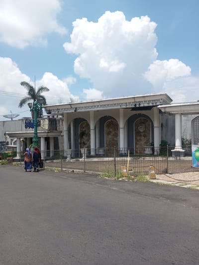 Two people walk on an asphalt street in front of a dilapidated white building with columns and arches, enclosed by a black metal fence. A palm tree and a green street lamp stand to the left of the building. The sky is blue with scattered white clouds. There is graffiti on a wall behind the building. The location is Xayabury District, Laos.