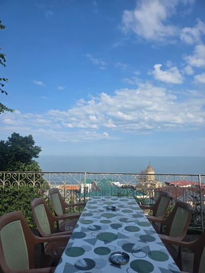 An outdoor seating area overlooks the Black Sea in Sarpi, Georgia. A table covered with a patterned tablecloth is set with two ashtrays. Four plastic chairs with woven seats surround the table. A decorative metal railing separates the seating area from the view. Beyond the railing, a lush green tree stands to the left, while buildings with red and green roofs are visible in the middle ground. A prominent building with a domed roof and a cross on top is situated further back. The calm, blue sea stretches to the horizon under a bright blue sky filled with scattered white clouds.