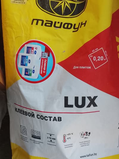 This is a close-up, eye-level shot of a bright yellow and white bag of adhesive. The bag has various text and graphics printed on it. At the top, there is text in Cyrillic and a logo. Below that, there is a circular graphic with three white bottles with red and blue labels inside. To the right of this, there is more Cyrillic text. In the lower half of the image, the word "LUX" is prominently displayed in large black letters. Below this, there is more Cyrillic text, along with several small icons depicting a cracked surface, a brick wall, a thermometer, and footprints. The bag appears to be standing upright. The background is blurred and out of focus. There is no indication of the location being in the city of Патоцкі сельскі Савет, Belarus.