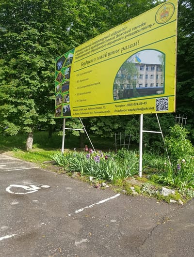 The image shows a yellow billboard with information about a college. The billboard has a college logo at the top and text that appears to be in Ukrainian. Below the text, there is a picture of a building with a Ukrainian flag. The billboard lists several specializations or departments. At the bottom of the billboard, there is an address, a phone number, and an email address. The address provided is: 37800, vul. Nebesnoi Sotni, 73, m. Khorol, Lubenskyi r-n., Poltavska obl. The billboard is situated outdoors, with trees and greenery in the background, suggesting a location in a park or near a college campus. The foreground shows a paved surface with white markings, possibly a parking lot with a designated disabled parking symbol. The overall scene is a bright, daytime outdoor setting.