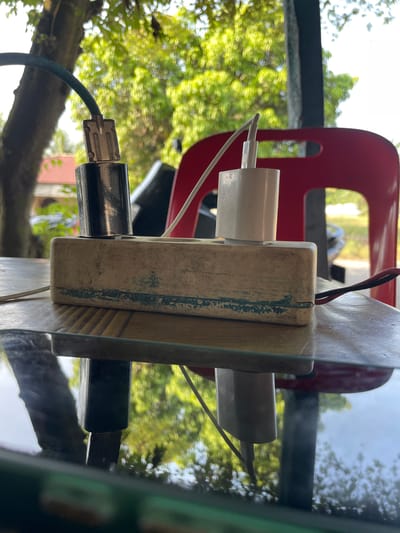 A close-up shot focuses on a power strip with two chargers plugged in, positioned on a reflective surface outdoors. A metallic charger with a blue cable is on the left, and a white charger is on the right. The power strip appears worn and discolored, with blue markings. In the background, out of focus, are lush green trees, a red chair, and a glimpse of a building and a car. The lighting suggests a bright day. The overall scene is captured from a low angle, emphasizing the chargers and power strip against the natural backdrop. This image was taken in Cotseutui, Indonesia.
