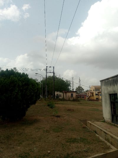 The scene is located in Alabata, Nigeria, depicting an outdoor environment under a partially cloudy sky. The ground consists of sparse, dry grass and exposed soil, with a slight uphill gradient towards the background. A dense, dark green shrub occupies the left foreground. Multiple overhead power lines extend across the upper and mid-sections, supported by a central utility pole with visible insulators and cross-arms, and at least one adjacent streetlight pole.

A light-grey building with a dark-framed window and an attached concrete retaining wall is partially visible in the right foreground. In the mid-ground, several smaller, single-story structures with light-colored, weathered walls are present. Further in the background, a multi-story building with yellow and orange sections and distinct architectural arches is visible on the right. Two lattice communication towers are positioned in the distant background, slightly left of the image center. No individuals or specific actions are observed within the frame.