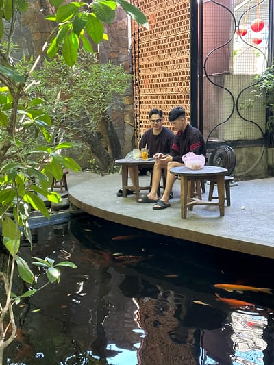 In a sunlit outdoor cafe setting in Da Nang, Vietnam, two young men sit at separate wooden tables situated near a koi pond. The pond's dark water reflects the surrounding environment, with several orange and gold koi fish visible swimming near the surface. The man on the left, wearing glasses and a dark t-shirt, has a drink on his table. The man on the right, wearing a black jacket with red stripes, is holding a mobile phone. A pink plastic bag rests on his table. The cafe features a textured brick wall in the background, a decorative metal screen, and lush green foliage. Small wooden stools are placed around the tables. The area appears to be a tranquil spot, offering a peaceful ambiance.