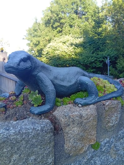 An artificial, dark blue lizard sculpture is positioned on a rocky outcrop covered with small succulents. The sculpture is detailed, with visible scales and limbs. It is resting on stones and dirt, with a wooden fence visible in the background to the left. Behind the fence, the environment transitions to lush green trees and foliage, suggesting an outdoor setting. The sky above is bright and slightly overcast. The location is the vicinity of the town of Sobótka, Poland.
