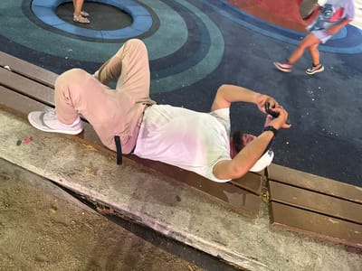 The image shows two people and a scene with multiple objects. A man wearing a white t-shirt, beige pants, a white cap, and white sneakers is lying on a wooden bench. He is holding a black smartphone in his hands and appears to be taking a picture or video. His gaze is directed towards the phone. To his left, a child is in motion, with their legs blurred, indicating movement. The child is wearing a light-colored t-shirt and shorts, and sneakers. They are on a colorful, circular play area with blue and red sections.
The setting appears to be an outdoor public space, possibly a park or playground, given the benches and the play area. The ground is a dark blue surface with concentric circles in shades of blue. The bench is made of dark brown wood and is placed next to a concrete area with dirt and scattered debris. The time of day is difficult to determine precisely, but the lighting suggests it is daytime. The overall mood seems casual and relaxed. No visible text is present in the image. The location is identified as Alcorcón, Spain.