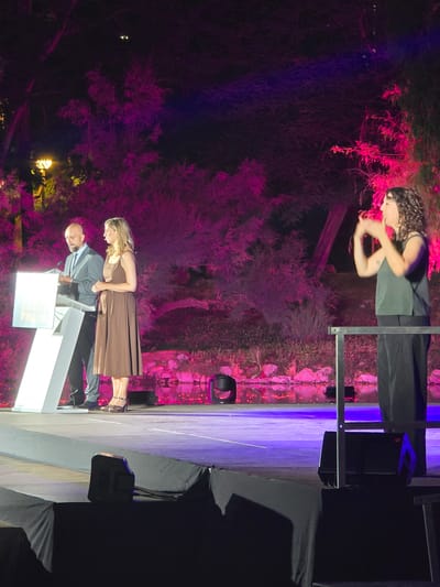 This is a medium shot of an outdoor event at night, likely in Cornellà de Llobregat, Spain. The stage is illuminated with pink and purple lights, casting a vibrant glow on the surroundings. The background features lush trees and a body of water.
On the left side of the stage, a man in a suit and tie stands behind a white podium, looking down at some papers. To his right, a woman in a brown dress stands beside him, looking towards the audience.
On the right side of the stage, a woman with curly hair is signing, her hands are in motion, and she is looking to her right. She is wearing a dark sleeveless top and black pants. A metal railing separates her from the main stage area.
In the foreground, parts of the stage equipment, including speakers and fabric coverings, are visible. The overall atmosphere suggests a formal event or presentation taking place outdoors after dark.