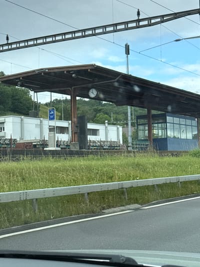 The image captures a sideways view of a railway environment, likely from a moving train or vehicle, looking towards a station platform in Zurzach, Switzerland. When viewed upright, the foreground on the left features intricate overhead catenary lines and support structures for an electric railway.

Central to the image is a train platform covered by a robust, dark-colored canopy. Suspended from the canopy's supports is a circular clock, which indicates the time as approximately 10:10. Beneath the canopy and extending behind it are several white, functional-looking station buildings. A prominent blue rectangular sign, typically used for platform identification, is visible in the midground, displaying a white left-pointing arrow and the number "1".

In the background, lush green hills covered in trees rise behind the station buildings. A modern blue building with large window panes is also discernible, reflecting parts of the cloudy sky. To the right of the platform infrastructure, a paved road or track runs parallel to a neatly maintained grassy verge, separated by a concrete barrier. The sky above is partially overcast with patches of blue visible. No people are apparent in the scene, which appears to depict a typical, quiet regional train station setting.