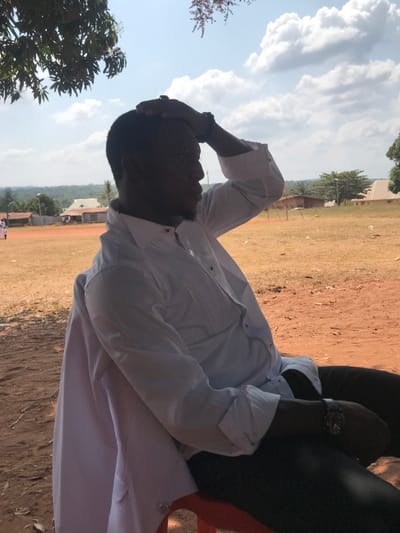 An adult male with dark skin and short dark hair is seated outdoors in Adum Afo, Nigeria. He wears a white long-sleeved shirt with vertical pleats on the chest and dark buttons, paired with dark trousers. His left hand is placed on the top of his head, fingers slightly splayed, while his right arm is bent with a dark watch on his wrist. A white garment is draped over the back of the red plastic chair he occupies. The subject's face is partially obscured by shadow, and he gazes towards the right side of the frame. The immediate ground consists of reddish-brown dirt with sparse, dry vegetation. In the mid-ground, an expansive, open field of similar reddish-brown earth and dry grass extends backward. In the background, a collection of single-story buildings with light-colored walls and dark roofs is visible, particularly to the left. Beyond these structures, a dense line of green trees marks the distant horizon. The sky above is bright blue with scattered white cumulus clouds, indicating strong sunlight, though the subject is partially shaded by an unseen overhead tree branch visible in the upper left. A few indistinct figures are visible on the field in the far left background.