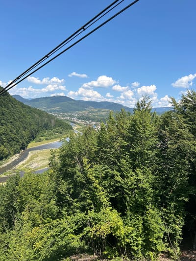 This is a photograph taken from an elevated viewpoint looking down at a town situated in a valley. The town, identified as Mizhhiria, Ukraine, is nestled between lush green mountains and bisected by a winding river. The sky above is a vibrant blue with scattered white, puffy clouds. In the foreground, the tops of numerous trees, dense with green leaves, obscure some of the view. Two dark cables, likely part of a cable car system, run diagonally across the upper portion of the image. The river appears shallow in places, with exposed gravel bars. The town itself consists of numerous buildings with red roofs, spread out along the riverbanks and up the hillsides. The overall scene depicts a scenic, natural landscape with a human settlement integrated into it.