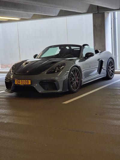 A gray Porsche sports car is parked in an indoor parking garage. The license plate, 'DR 0109', is visible. The car is parked between a white painted wall and a marked parking line. The 'AUTOPOLIS' is labeled on the license plate. The location of the scene is Sanem, Luxembourg.
