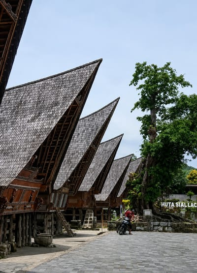 The image captures a row of traditional houses with distinct saddle-shaped roofs in Ambarita, Indonesia. The roofs are covered with grey, overlapping shingles. The houses are elevated on stilts and feature ornate wooden carvings on their facades. A paved walkway runs in front of the houses. In the foreground, a person wearing a red shirt and dark pants is standing next to a motorcycle. To the right of the houses, a large tree with dense green foliage stands prominently. In the background, stone structures are visible, with a sign that reads "HUTA SIAL LAC." The sky is clear and blue.