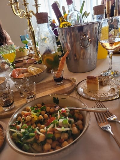 The image displays a dining table set indoors in Favões, Portugal, against a backdrop of white curtains. A white tablecloth covers the table, laden with various food and beverage items. A silver ice bucket, containing two bottles (one with a gold label, another clear), stands prominently. Adjacent to it, a clear glass carafe with a cork stopper holds a lemon and mint-infused drink. To the left, a tall, ornate gold candelabra is partially visible. In the foreground, a ceramic bowl containing a chickpea salad with chopped peppers, onions, and greens holds a silver spoon. Another bowl to its left contains peeled shrimp and lemon slices. A shot glass contains a fried shrimp tempura in a dark liquid. On the right, a stemmed glass holds a golden beverage, and a decorative glass plate features a slice of cake and a smaller pastry. Multiple silver forks are arranged on the tablecloth to the right of the chickpea salad. Other objects include a green textured glass with orange liquid, appetizers on skewers on a wooden board, and multiple shot glasses on a wooden tray. A person's arm in a light brown sleeve is visible on the far left, and the upper head of another individual is seen in the upper right.