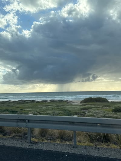 The image presents a coastal scene near Tirat HaCarmel, Israel. Dark storm clouds occupy the left portion of the frame, transitioning into a brighter, sunlit area above the sea. The sea itself has visible waves. A grassy embankment and a metal guardrail are situated on the right, adjacent to an asphalt road. No people or subjects are visible. The general environment suggests a stormy coastal region near a road.