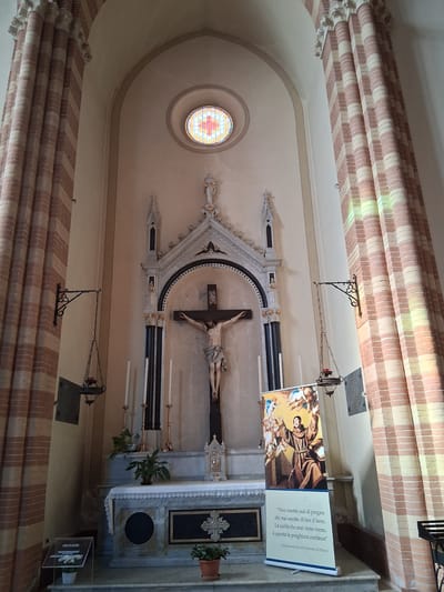 The image depicts the interior of a church in Padua, Italy, specifically the altar area. A crucifix with a statue of Jesus is centrally positioned above an altar, which is adorned with a white cloth and various decorative elements. To the left of the altar, there are several tall candles and plants. To the right, a banner featuring an image of Saint Anthony of Padua with a quote in Italian is displayed. The church architecture includes large, fluted columns with alternating red and cream stripes, and a stained-glass window at the top of the apse. Hanging from the ceiling are ornate lanterns. The overall lighting suggests a natural light source, with some colored light filtering through the stained glass. The scene is static and appears to be captured during the daytime.