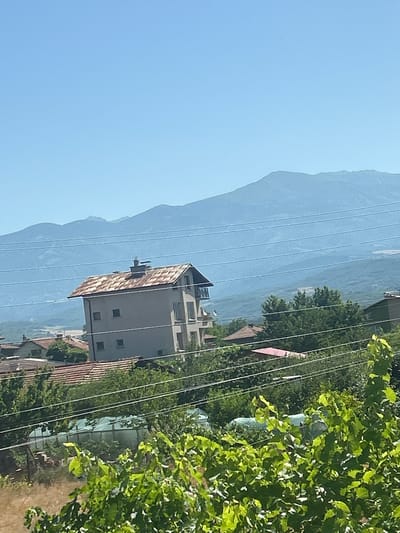 A multi-story building with a rusty, sloped roof stands in the middle ground of this outdoor, daytime photograph. The building is light-colored with several windows and a chimney. The scene appears to be a residential area in Boboshevo, Bulgaria, with other houses and lush green trees visible in the foreground and mid-ground. In the background, distant mountains are shrouded in a light haze under a clear blue sky. Power lines are strung across the scene, crossing in front of the main building and the mountains. The overall impression is a rural or semi-rural landscape.