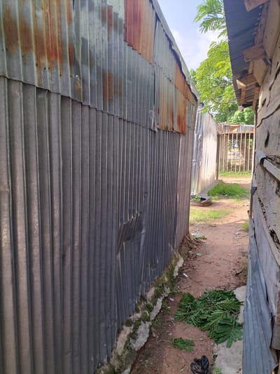The image depicts a narrow outdoor pathway between two structures, likely in Jauro Bamvo, Nigeria. On the left, a corrugated metal wall with visible rust streaks and some damage runs along the path. On the right, a section of a wooden structure with peeling material is visible, also bordering the pathway. The ground is a reddish-brown dirt path, with some patches of green grass and scattered leaves. In the background, a metal gate with vertical bars can be seen, beyond which is more greenery and trees. The overall scene suggests an informal or utilitarian setting.