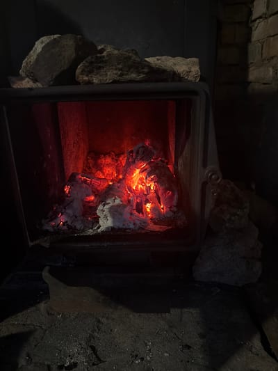The image shows the inside of a wood-burning stove, likely in a rustic setting in Argamak, Russia. The stove's metal door is open, revealing a pile of glowing embers and ash. The embers are a vibrant orange and red, indicating that the fire is still active, though diminishing. White and grey ash is mixed with the embers, creating a textured, almost sculpted appearance.
The interior of the stove appears to be made of metal, with some heat-resistant bricks or stones visible around the edges. The area surrounding the stove is dark and appears to be made of rough, textured material, possibly stone or concrete. Two large, grey stones are positioned on the left side of the stove opening, acting as a natural surround or support. Above the stove, dark brickwork is visible, suggesting it is built into a chimney structure.
The lighting in the image is dim, with the primary light source being the glow from the embers. This creates a dramatic contrast between the bright, hot center and the dark surroundings. The overall impression is one of warmth and residual heat from a recently burned fire. There is no indication of the time of day or weather, but the scene evokes a sense of coziness and the comfort associated with a fire on a cold day, common in rural Russian locations. No text is visible in the image.