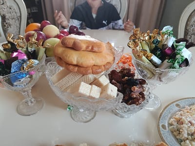 A medium shot captures a table laden with sweets and fruits, likely for a gathering or celebration. In the foreground, a crystal bowl holds an assortment of foil-wrapped chocolates and candies. To the left of this, another crystal bowl contains a mix of fruits including apples, peaches, and oranges. In the center, a stack of golden-brown cookies, possibly shortbread or bisquits, sits atop a layer of wafer cookies, also presented in a crystal bowl. To the right of the cookies, a bowl filled with dates and nuts is visible. Further to the right, a white plate with a decorative border holds a serving of salad, likely a potato salad or Olivier salad, with visible chunks of vegetables and mayonnaise. In the background, a person, whose face is partially obscured, is seated at a table, with their hands gesturing. The setting appears to be indoors, with a hint of patterned curtains and furniture visible. The lighting is bright, suggesting daylight or well-lit interior conditions. The overall scene suggests a festive or familial occasion, with an emphasis on sweets and shared food. The location is Komsomol'skoye, Kyrgyzstan.