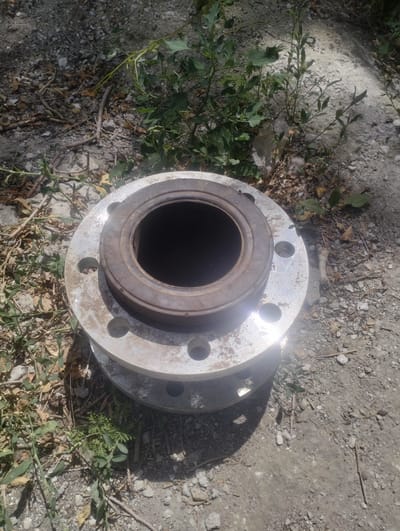 The image shows a close-up, top-down view of two large, circular metal flanges stacked on top of each other. The flanges are silver and have a series of bolt holes around their circumference. The top flange has a raised, darker metal ring in its center, creating a hollow opening. The objects are resting on a surface of dirt, gravel, and dried leaves, with some sparse green vegetation growing around them. The lighting suggests it is daytime, with a bright sun casting shadows. The scene appears to be outdoors in a natural, possibly unkempt, environment. There is no visible text in the image. The context of Qarabulaq, Kazakhstan, suggests a location that could be industrial or rural. The objects themselves appear to be industrial components, possibly part of a pipeline or machinery. The overall impression is one of discarded or stored industrial equipment in an outdoor setting.