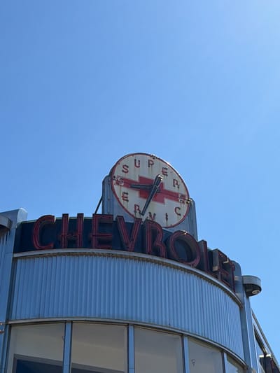 The image shows the exterior of the former Chevrolet dealership located in Rochester, New York. The iconic neon sign is visible, featuring the word "CHEVROLET" in large red letters. Above the sign is a circular clock with the words "SUPER SERVICE" around the Chevrolet logo. The building has a curved facade with corrugated metal siding and large windows. The sky is a clear blue.