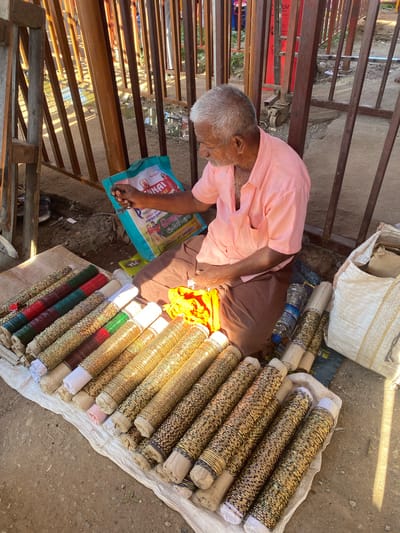 An elderly Indian man with gray hair and a mustache, wearing a light pink collared shirt and brown pants, sits on the ground next to a display of colorful bangles in Chinnambedu, India. He is holding a blue shopping bag with Tamil writing and images of people. The bangles are arranged in rows on a white cloth, with some wrapped around cylindrical cardboard tubes. Other items visible include a bright orange cloth, a plastic water bottle, and a large sack. The scene appears to be an outdoor market, with metal bars and a dirt ground. The lighting suggests it is daytime, with sunlight casting shadows.