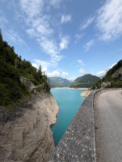 A wide-angle, eye-level, outdoor shot captures a scenic view of a turquoise lake in Ampezzo, Italy, bordered by lush green mountains and dense pine forests. The lake's water is calm, with a few buoys visible. The foreground features a concrete dam structure, with a paved road curving around its edge. The road has a wooden railing separating it from the drop-off. On the left side of the image, a steep, rocky cliff face descends towards the lake, showcasing weathered concrete structures and remnants of past construction. The sky above is a vibrant blue, dotted with scattered white clouds, indicating a clear, sunny day. The overall impression is one of natural beauty and human engineering in harmony.