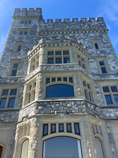 The image shows a low-angle, full-frame shot of the exterior of a large stone building, possibly a castle or large estate, under a clear blue sky with sparse clouds. The building is constructed of irregular grey and white stones, with lighter colored stone detailing around windows and architectural features. The structure has multiple stories with numerous windows of various shapes and sizes. Some windows are rectangular with dark frames, while others are arched. There are decorative battlements along the top of the building. A red and white flag is visible at the very top of the highest tower. The architecture suggests a gothic or medieval influence. This building is Hatley Castle, located at Royal Roads University in Victoria, British Columbia, Canada. It is not in the city of Langford, Canada.