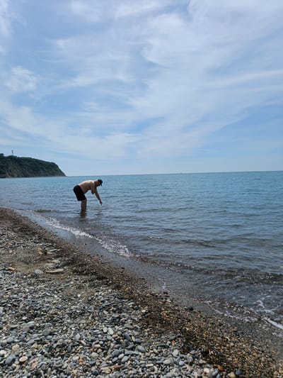 The image presents a coastal scene in Krinitsa, Russia, under a partly cloudy sky. In the midground, a lone adult male is standing in the shallow, calm seawater, bent at the waist with his right hand submerged just below the surface. He is shirtless, wearing dark shorts and a dark baseball cap. The foreground reveals a pebble beach composed of numerous rounded stones in shades of grey, brown, and white, extending from the bottom of the frame to the water's edge. Small waves gently break on the shore, creating minor foam and glints of light on the water's surface. The sea, exhibiting a blue-grey hue, stretches towards the horizon with a placid surface. On the left side of the frame, a verdant, forested hill or cliff slopes down to meet the water. A tall, slender tower-like structure is visible atop this landmass. The sky occupies the upper half of the image, featuring scattered, wispy white clouds against a pale blue backdrop.