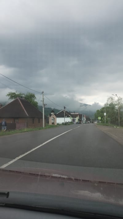 The image shows a view from inside a vehicle, looking out onto a road in Yasinia, Ukraine. The road is asphalt, marked with a white line dividing the lanes. To the left of the road are buildings, trees, and utility poles, partially obscured by a cloudy sky. A mountain is visible in the distance, shrouded in mist. The right side of the image shows the interior of the vehicle, including part of the dashboard and window frame.