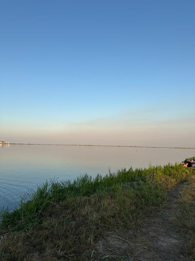 In Serhiivka, Ukraine, a tranquil scene unfolds by the water. The image captures a wide expanse of calm water reflecting the clear blue sky. The horizon is marked by a distant, low-lying shoreline. In the foreground, a grassy embankment slopes down to the water's edge. The grass is lush and green in some areas, transitioning to dry, brown vegetation in others. A dirt path is visible on the embankment, leading towards the right side of the frame.
On the right side of the image, near the top of the embankment, two individuals are seated on chairs. They appear to be relaxed, with one person looking towards the water. Several items, including bags and possibly blankets, are arranged around them. A small orange buoy is visible in the water, further out from the shore. In the distance, along the left-hand horizon, a building with a red roof is faintly discernible. The overall atmosphere is peaceful and serene, suggesting a leisurely day by the water.