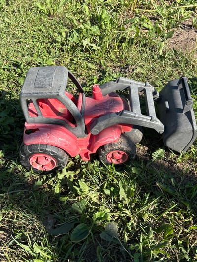 A red toy tractor with a gray loader is lying on its side in a grassy area. The tractor is plastic and appears to be old and somewhat damaged, with some cracks visible on the body. The loader is raised and angled forward. The ground is covered in green grass and some dry dirt. The image is taken from a slightly elevated angle, looking down on the toy. There are no people or animals visible in the image. The lighting suggests it is daytime, likely outdoors. The provided location of Новый Кинер, Russia, does not offer specific environmental context visible in the image itself.