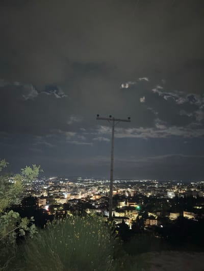 The image shows an aerial view of a city at night, potentially Xanthi, Greece. City lights illuminate buildings and streets. In the foreground, there is an electrical pole. The sky is cloudy. The shot is taken from an elevated position, with vegetation visible at the bottom and right of the frame.