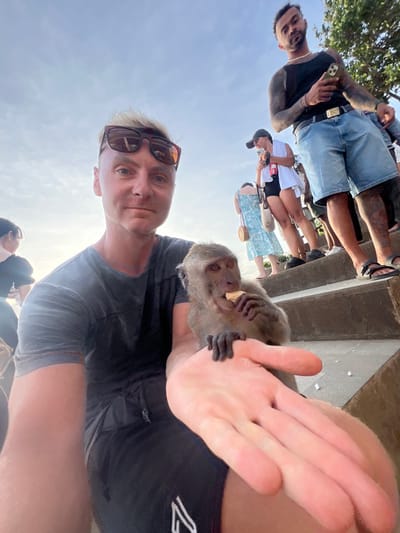 A male individual is depicted in a selfie-style photograph taken outdoors in Bunga Mekar, Indonesia. He has blond hair, sunglasses resting on his head, and wears a dark grey t-shirt and black shorts. A small, brown-furred monkey is situated on his left arm, actively consuming a light-colored, flat food item. The monkey's left paw is placed on the man's extended right hand, which is likely holding the camera. In the background, several people are observed on concrete steps ascending towards the right. One male, with dark hair and visible arm tattoos, wears a black tank top, blue denim shorts, and sandals, holding a mobile phone and looking towards the camera. Behind him, a female wearing a white top, dark shorts, and a baseball cap is also holding a phone. Another female in a light blue patterned dress and a circular woven bag is partially visible further back. Green foliage is present on the right side of the frame, and the scene is illuminated by natural daylight under a bright sky.