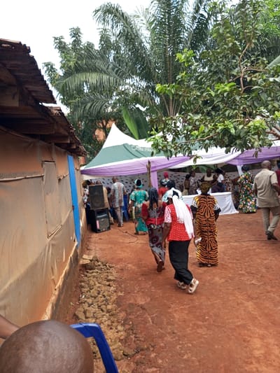 The image captures an outdoor gathering in Oko Town, Nigeria, characterized by a prominent red earth ground surface. To the left, a building with a corrugated metal roof and light-colored, possibly fabric-covered, walls is visible, featuring a vertical blue section. Adjacent to this wall, two black audio speakers are stacked, with associated wiring on the ground. A line of loose rocks and soil runs along the base of the building. In the mid-ground, a white and green tent with purple decorative fabric is erected, sheltering a group of individuals. Numerous people, predominantly adults, are dressed in vibrant, patterned traditional attire, with several individuals seen walking towards or congregating under the tent; a white table is also visible within the tent area. Specific individuals include a woman wearing a red patterned top, black trousers, white sandals, and a white headscarf; another woman in a patterned dress; and a third woman in a long, tiger-striped dress. The background features lush tropical vegetation, including tall palm trees and various green leafy trees. The upper portion of a human head and part of a blue plastic chair are visible in the bottom-left foreground.