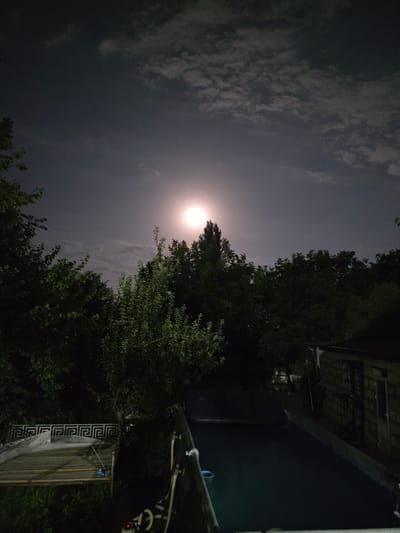 The image depicts a nighttime scene in Yalangach, Uzbekistan, illuminated by a bright full moon. The moon is prominently visible in the upper center of the frame, surrounded by scattered clouds. A dense line of dark green trees forms a silhouetted backdrop across the mid-ground. In the foreground, to the left, is a wooden structure covered with a light-colored tarp, featuring a decorative geometric railing. To the right, a dark, flat surface, possibly a pool or pond, occupies the lower portion of the image, leading towards a small, brick building with a closed door and window. The overall atmosphere is serene and dark, typical of a rural or suburban night.