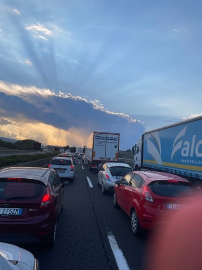 The image displays a multi-lane highway experiencing a traffic jam at sunset near Paliano, Italy. The sky is dominated by dark, horizontally stratified clouds in the mid-frame, from which prominent crepuscular rays emanate, fanning across the upper sky. Golden light from the low sun is visible beneath the clouds. Green, rolling hills form the distant background on the left side of the highway.

The roadway is densely packed with stationary or very slow-moving vehicles. In the immediate foreground, the rear of a dark red Ford Fiesta hatchback (Italian license plate FC 275ZM) is visible on the left, with its taillights illuminated. A portion of a white car is visible at the bottom left. In the adjacent lane to the right, a red Ford Fiesta hatchback (Italian license plate FF 443RY) is present, with its lower right obscured by a blurred, reddish object. Ahead of this vehicle is a silver Lancia Ypsilon (Italian license plate EK 13XX), followed by a white hatchback (Italian license plate EJ 825CJ).

Further down the congested highway, a white semi-trailer truck marked "Rosso Trasporti" is visible in the middle lane, followed by numerous other cars. In the rightmost lane, a large blue semi-trailer truck with "Falco Broker & Log" on its side is prominent. The asphalt features clear white dashed lane markings. No individuals are discernible.