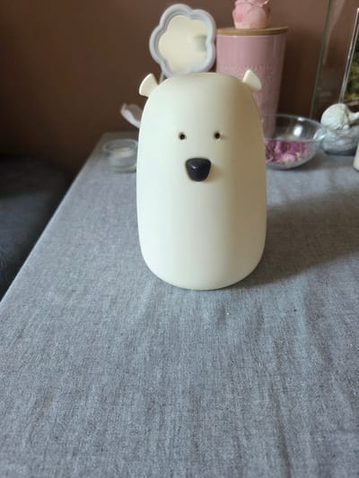 A white, stylized bear-shaped object is centered on a grey, textured tablecloth. The object has two small, black dots for eyes and a black, triangular nose. Two small, white ears protrude from the top of its head. The background is blurred, showing a brown wall, a pink cylindrical container with a pink flower decoration on top, a clear glass bowl containing pink dried petals, and other decorative items. The scene appears to be indoors, on a table. The location in Brzeg, Poland, is not ascertainable from the provided image.