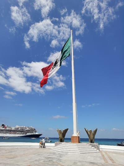 The image shows the Mexican flag flying high on a flagpole in front of the Cozumel Cruise Terminal, located in Cozumel, Mexico (latitude: 20.511, longitude: -86.951). The flag is waving gently in the breeze against a bright blue sky with white clouds. Two bronze sculptures of eagles are positioned on either side of the flagpole, and there are two people standing near the sculptures. A large cruise ship is visible in the background, docked at the terminal. The scene is a bright, sunny day with clear, blue water in the background.