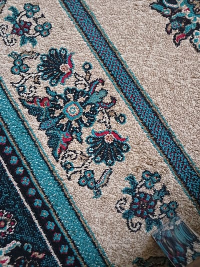 The image shows a close-up view of a carpet with an intricate floral pattern. The carpet has a predominantly beige background with diagonal bands of alternating dark blue and turquoise woven with red accents. The floral motifs are stylized, featuring dark blue and teal petals with hints of red and pink. The texture of the carpet appears plush and textured. In the bottom right corner, a portion of a cleaning tool, possibly a carpet cleaner attachment, is visible, suggesting an action of cleaning or maintenance. The scene is indoors, indicated by the lighting and the material of the carpet. The image itself does not provide any specific information about its location in the city of Guba, Azerbaijan, or any details about people or specific events.