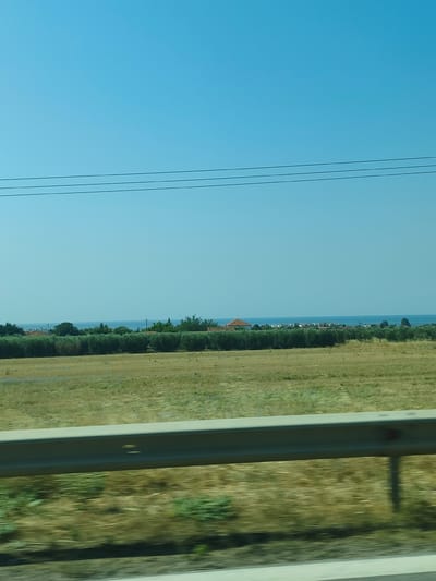 The image captures a rural landscape under a clear blue sky. In the foreground, a metal guardrail runs horizontally across the bottom of the frame, partially obscuring a blurry field of dry grass and green vegetation. Beyond the field, a dense line of olive trees with terracotta-roofed buildings peeking through them stretches across the middle ground. In the distance, the calm, blue sea is visible, meeting the horizon. Three dark, thin power lines are strung across the upper portion of the sky. The scene appears to be located in a coastal area near Σωζόπολη, Greece.