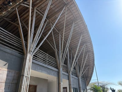 The photograph captures the exterior of a building constructed with bamboo in Nusa Penida, Indonesia. The building features a distinctive curved roof made of bamboo poles and thatch, supported by an intricate network of diagonal bamboo columns that fan out towards the roofline. A concrete facade forms the lower levels of the structure, with horizontal bamboo railings visible on an upper balcony. Sunlight casts shadows on the building's surfaces, indicating a bright, sunny day. The sky is a clear, pale blue. In the distance, foliage and a dark silhouette of a statue are visible. The scene is static, with no people or discernible activity present.
