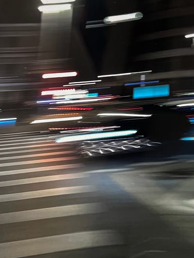 The image depicts a motion-blurred night scene in what appears to be an urban environment. Horizontal streaks of light, characteristic of moving vehicles or illuminated signage at night, dominate the composition. The foreground shows what seems to be a crosswalk with painted white lines, some illuminated by the vehicle lights. In the background, various colored lights, including white, red, and blue, are streaked horizontally, suggesting traffic and possibly advertising or building lights. The dark areas suggest a night sky or unlit portions of buildings and roads. The overall impression is of movement and the ephemeral nature of urban light at night. There is no specific visual evidence within the image to confirm its location in Genemuiden, Netherlands.