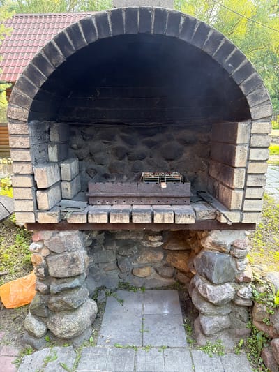 A rustic, outdoor brick barbecue oven, constructed with a stone base and a brick archway, sits in a yard in Novy Gorodok, Russia. The oven's interior is dark and sooty, with a metal grill rack positioned inside. Loose bricks are stacked to the left of the grill, and a metal spatula lies on the shelf in front of the oven. The ground around the barbecue is a mix of grass and paving stones, with an orange plastic bag visible to the left. In the background, a portion of a wooden house and a red-tiled roof can be seen, surrounded by green trees.