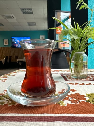 The image depicts a close-up view of a glass of dark red tea on a table covered with a tablecloth. The tablecloth features a pattern of autumn leaves in shades of orange, brown, and green, with horizontal stripes of brown and cream. The tea is served in a traditional tulip-shaped glass and sits on a matching glass saucer. To the right of the tea, a small green plant in a clear glass jar is visible, its roots submerged in water. In the background, the interior of a cafe or restaurant is partially visible. The walls are painted teal and black. A flat-screen television is mounted on the teal wall, displaying a vibrant image of a city street at night. There are framed pictures on the wall. Tables and chairs are set up in the dining area. The ceiling has modular lighting fixtures. The scene suggests a relaxed atmosphere within a commercial establishment.
While the image is detailed, there is no specific visual information that allows for the precise identification of the location as Sosnowiec, Poland.