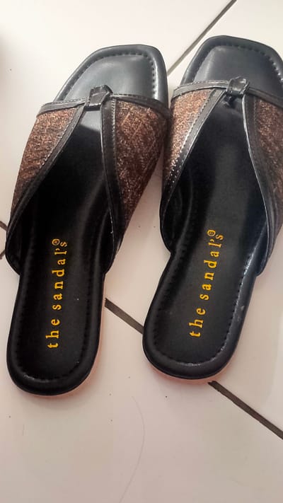 Two black sandals, each with a brown patterned strap, are laying on a white tiled floor. The sandals are labeled "the sandals" on the sole in yellow lettering. The sandals are likely located inside a residence in Bojong Gede, Indonesia. The floor is tiled and there are small specks of dirt on the tiles. The sandals are laying flat on the floor. They are not being worn and are not being interacted with. The lighting appears to be artificial and overhead. The image is taken from an overhead angle. The background is a white tiled wall with the tiles visible at the edges of the image.