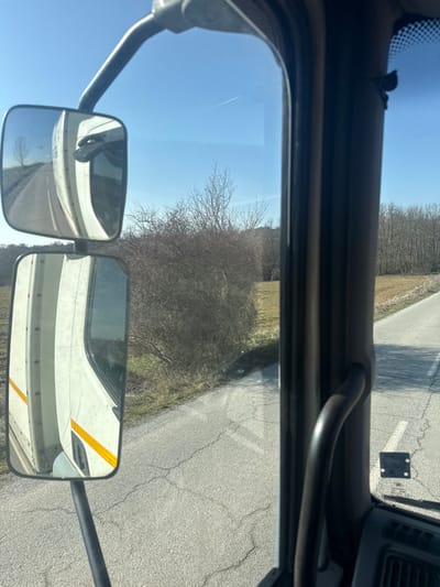 The image depicts a static view from the interior of a vehicle, looking out its right-side window onto a rural road and landscape in Begunovtsi, Bulgaria. The immediate foreground on the right features a dark, curved grab handle attached to a vehicle pillar. To the left, two rectangular side mirrors are positioned, reflecting the white exterior of the vehicle's cabin, which has a distinct horizontal yellow reflective stripe. The mirrors also reflect a segment of a paved road receding into the distance, with a solitary bare tree visible further back.
Outside the vehicle, a grey asphalt road, displaying numerous surface cracks, runs generally parallel to the vehicle's orientation. Adjacent to the road is a narrow strip of dry ground and sparse vegetation. Beyond this, a dense line of bare deciduous bushes and trees forms a natural boundary, separating the road from an expansive, light-brown field of dry grass or stubble. In the far background, a dense forest of leafless trees covers a gently rising terrain. The sky is clear and bright blue, traversed by a single, thin white contrail from an aircraft. The scene is illuminated by direct sunlight, and no people or active subjects are visible.